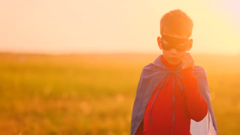 Ein-Als-Superheld-Verkleideter-Junge-Steht-Mit-Maske-Und-Rotem-Umhang-Da-Und-Beobachtet-Im-Sommer-Den-Sonnenuntergang-Auf-Einem-Feld.-In-Einer-Sommernacht-Träumt-Der-Junge-Von-Heldentaten-Und-Comics