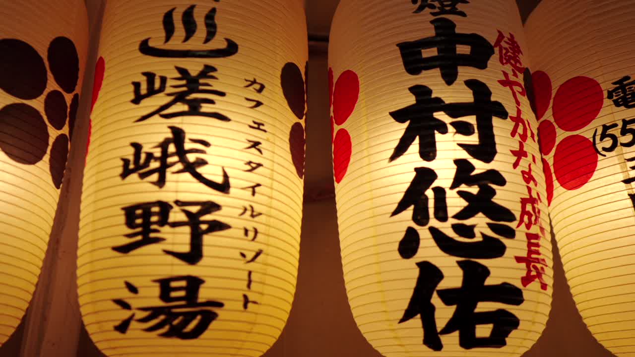 Primo piano panoramico di lanterne tradizionali giapponesi illuminate appese al Santuario Nishiki Tenmangu a Kyoto, Giappone, che mostrano vari caratteri Kanji e disegni
