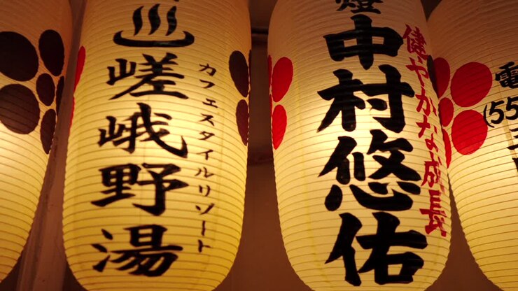 Primo piano panoramico di lanterne tradizionali giapponesi illuminate appese al Santuario Nishiki Tenmangu a Kyoto, Giappone, che mostrano vari caratteri Kanji e disegni