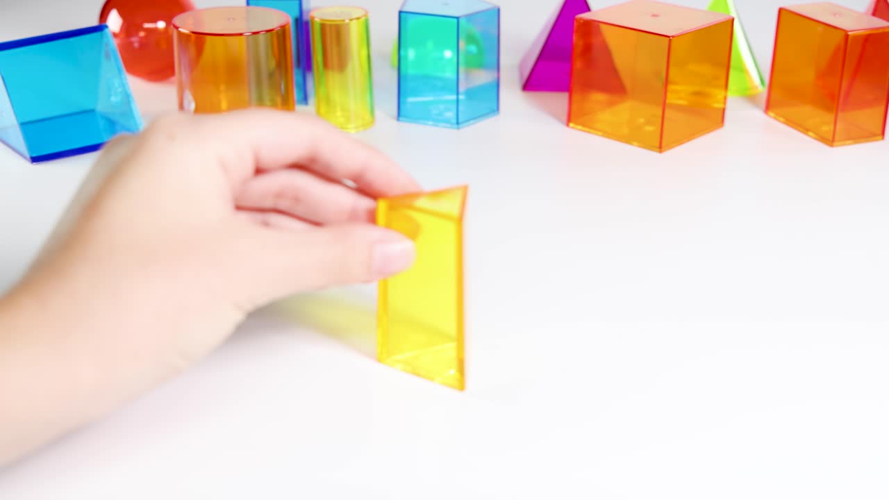 A hand manipulates vibrant 3D geometric shapes on a white surface, demonstrating movement and interaction in a bright setting