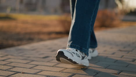 a close-up image of a person wearing jeans and white shoes walking slowly on a paved path in a park. a sense of calmness mixed with sadness, as the deliberate, slow steps reflect a subdued mood