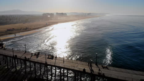 beautiful sunrise with shimmering pacific ocean aerial 4k drone pull back from waves and sun to reveal huntington beach pier and surfers in early morning