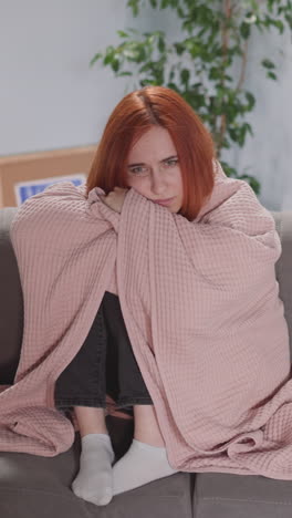 redhead woman watches interesting serial on tv covering with warm pink plaid. pensive female plunged into story of characters sits on sofa in living room