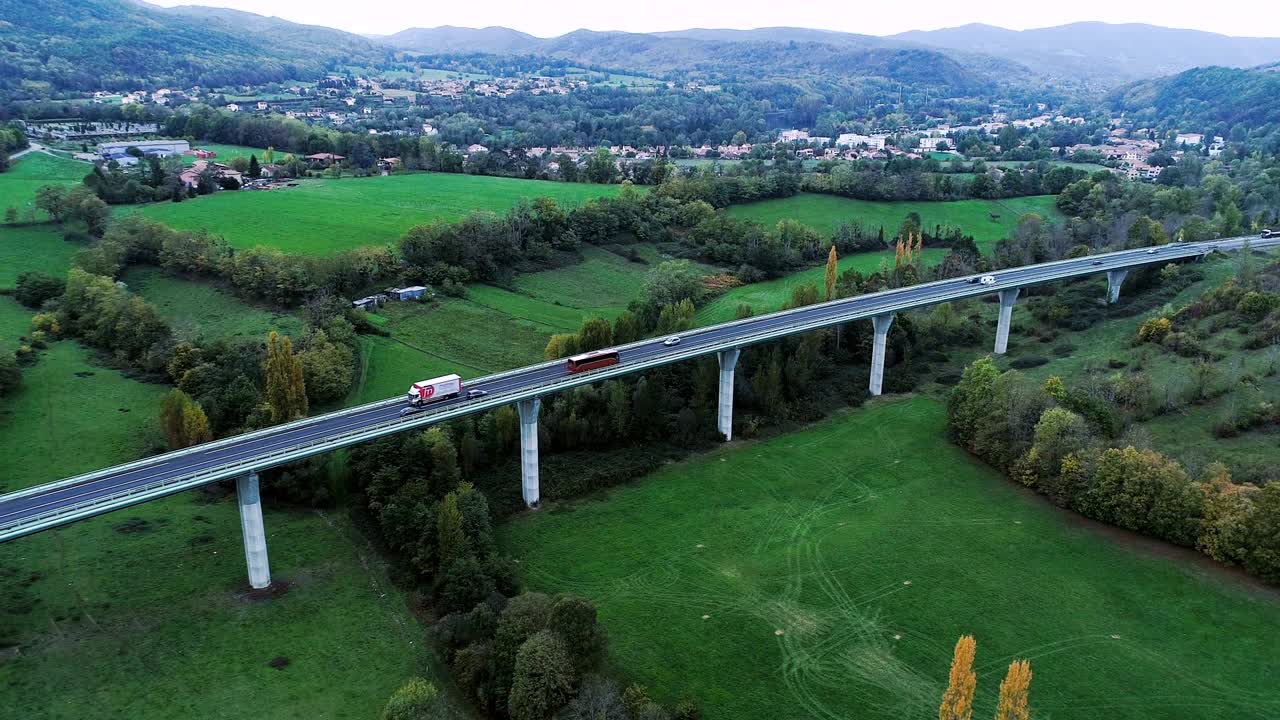 프랑스의 한 다리 위의 고속도로 위의 공중 풍경: 도로 주변의 잔디과 나무들, 빛 아래 도로를 달리는 자동차와 트럭들.