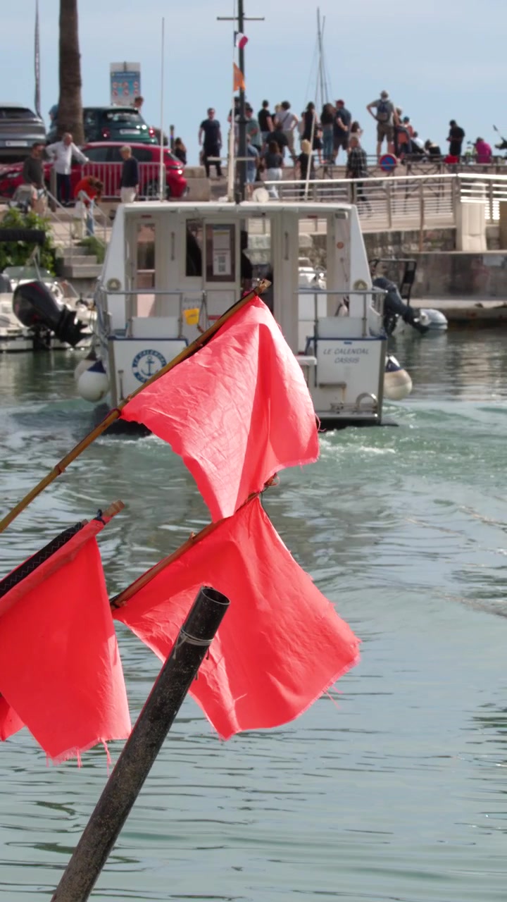 Bandiere rosse sventolano in primo piano mentre una barca turistica parte dal porto di Cassis, Francia.