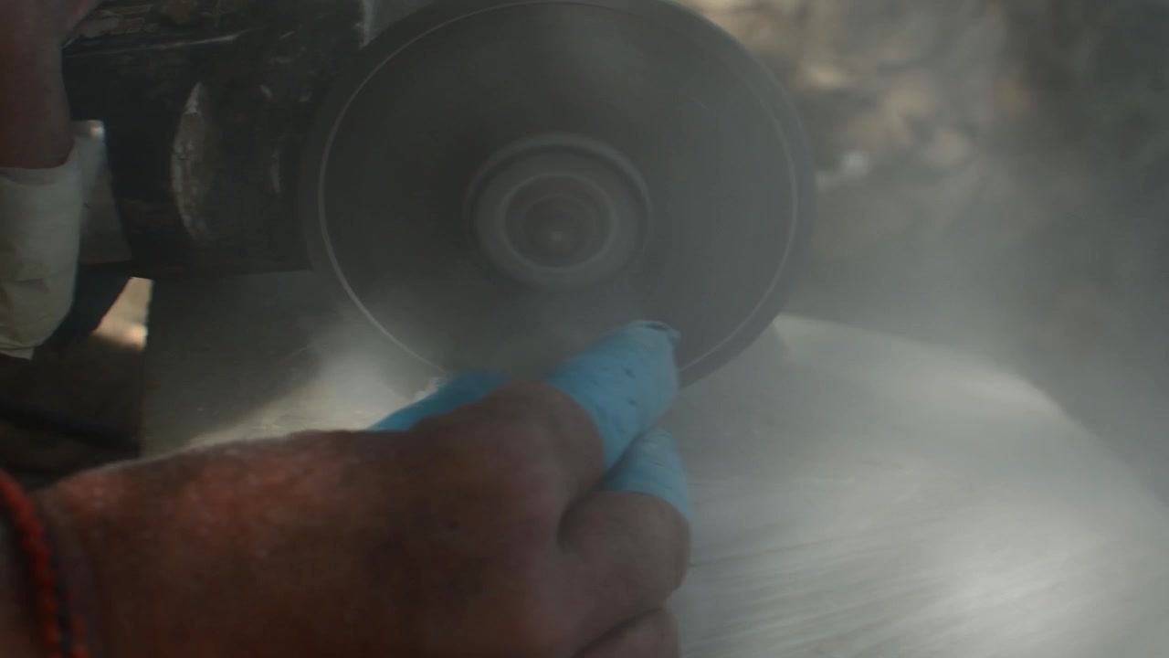 Gros plan sur les mains gantées d'un ouvrier utilisant un outil électrique, créant de la poussière en taillant la pierre dans un atelier.