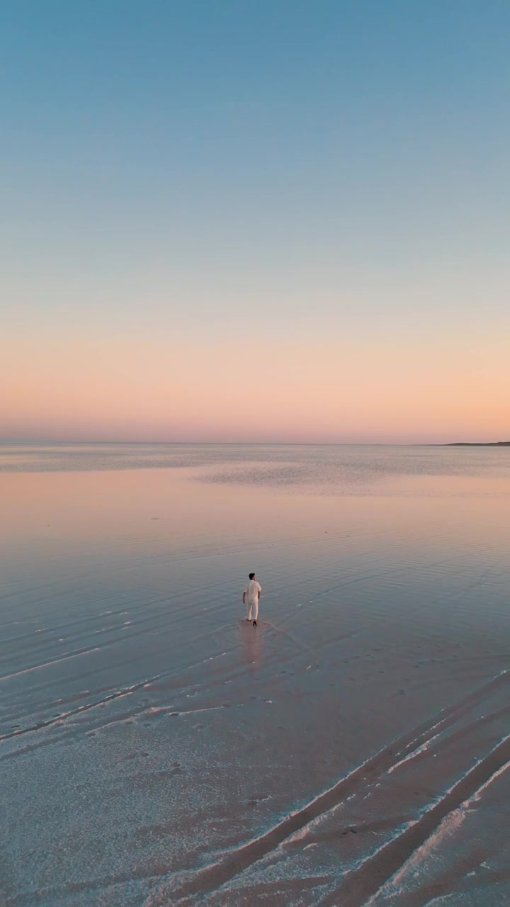 Video verticale in stile social media di un uomo che cammina su acqua calma e riflettente sotto un cielo al tramonto dai colori pastello con un'atmosfera tranquilla e minimalista.