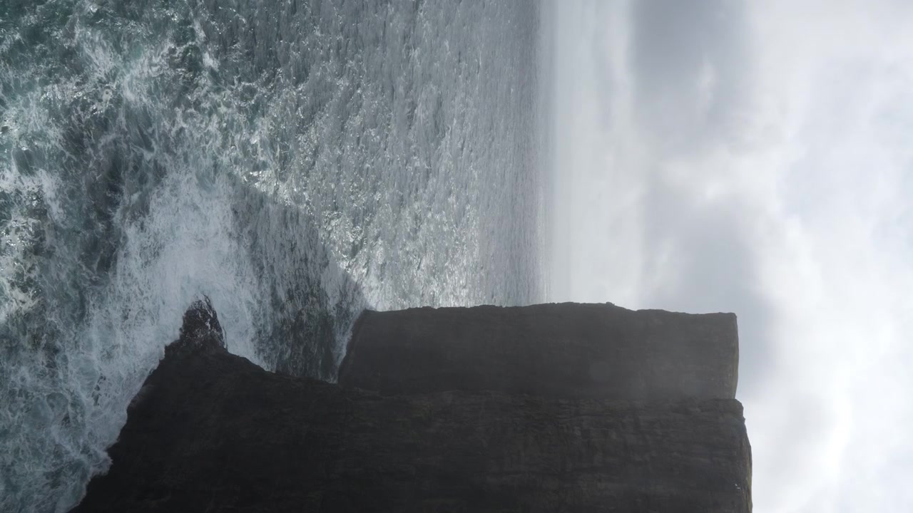 Vertical shot of cliff casting shadow on sea water on partly cloudy day, Faroe Islands