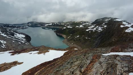 Flying-away-from-a-lake-with-blue-glacier-water,-revealing-the-landscape-with-rocks-and-a-snow-covered-glacier