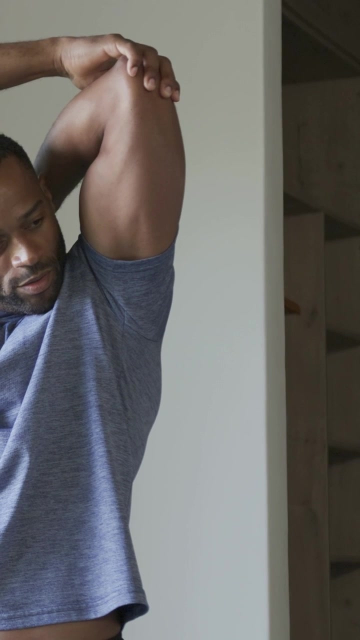 Vertical video: Raising right arm, man stretching triceps at home near shelving, in gray tee