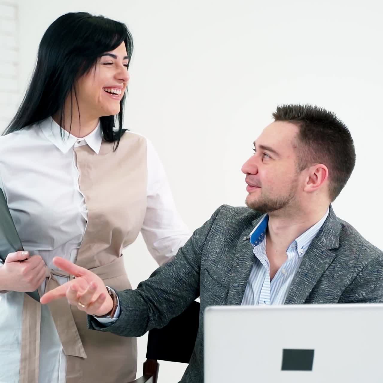 Businessman talking to a young smiling secretary. Handsome man in suit sitting with a laptop in his hands and talking to a beautiful woman with a folder.