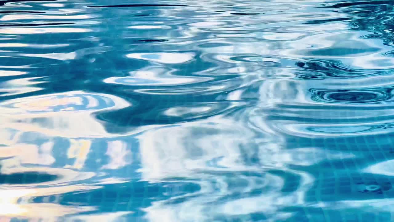 Clear water ripples gently in a swimming pool, its surface reflecting sunlight and revealing the blue mosaic tiles below