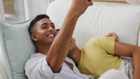 Smiling-mixed-race-gay-male-couple-hugging-lying-on-sofa-taking-selfie