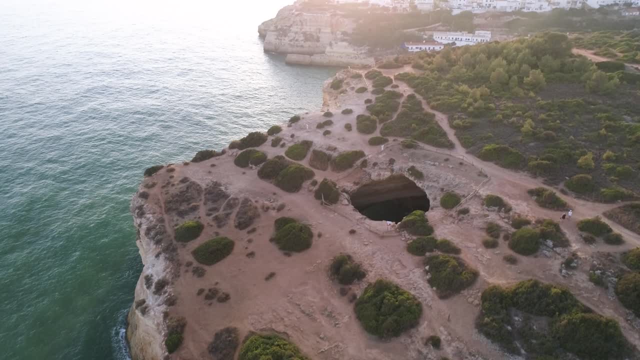 Benagil caves and the village in the background. Drone shot rising and tilting to reveal a tourist boat entering the caves.