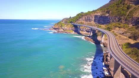 Aerial-footage-of-a-stunning-long-curvy-Bridge-next-to-the-sea-hightlights-the-Grand-Pacific-Drive-with-south-pacific-ocean-and-blue-sky-view-on-a-bright-sunny-day,-Sydney,-Australia