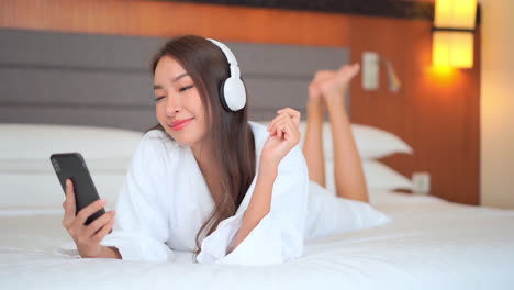 happy pretty asian woman watching phone and listen to music while lying on the hotel bed wearing a white dressing gown, smiling front view