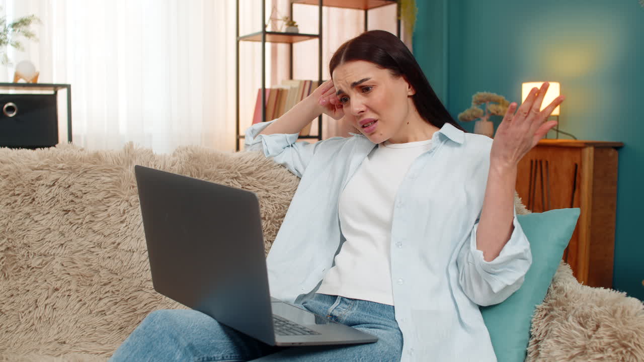 Young woman on sofa looking dissatisfied at laptop screen leaning head on hand tired and upset