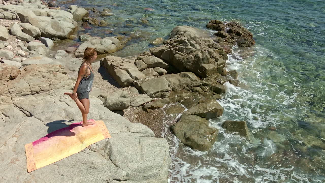 mujer de pie sobre una roca junto al mar practicando yoga en un día soleado en cerdeña, italia - toma aérea de drones
