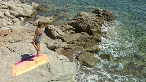 Woman-Standing-On-A-Rock-By-The-Sea-Practicing-Yoga-On-A-Sunny-Day-In-Sardinia,-Italy---aerial-drone-shot