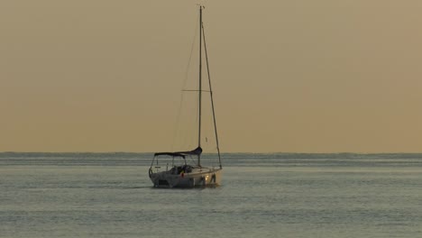 Sailboat-at-dawn-on-calm-ocean-with-sails-furled,-heading-out-to-sea