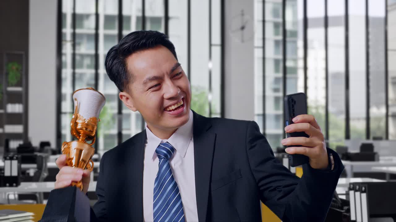 Close Up Of Asian Business Man In A Suit And Tie With A Gold Medal Showing A Gold Trophy To The Video Call On Smartphone In Office