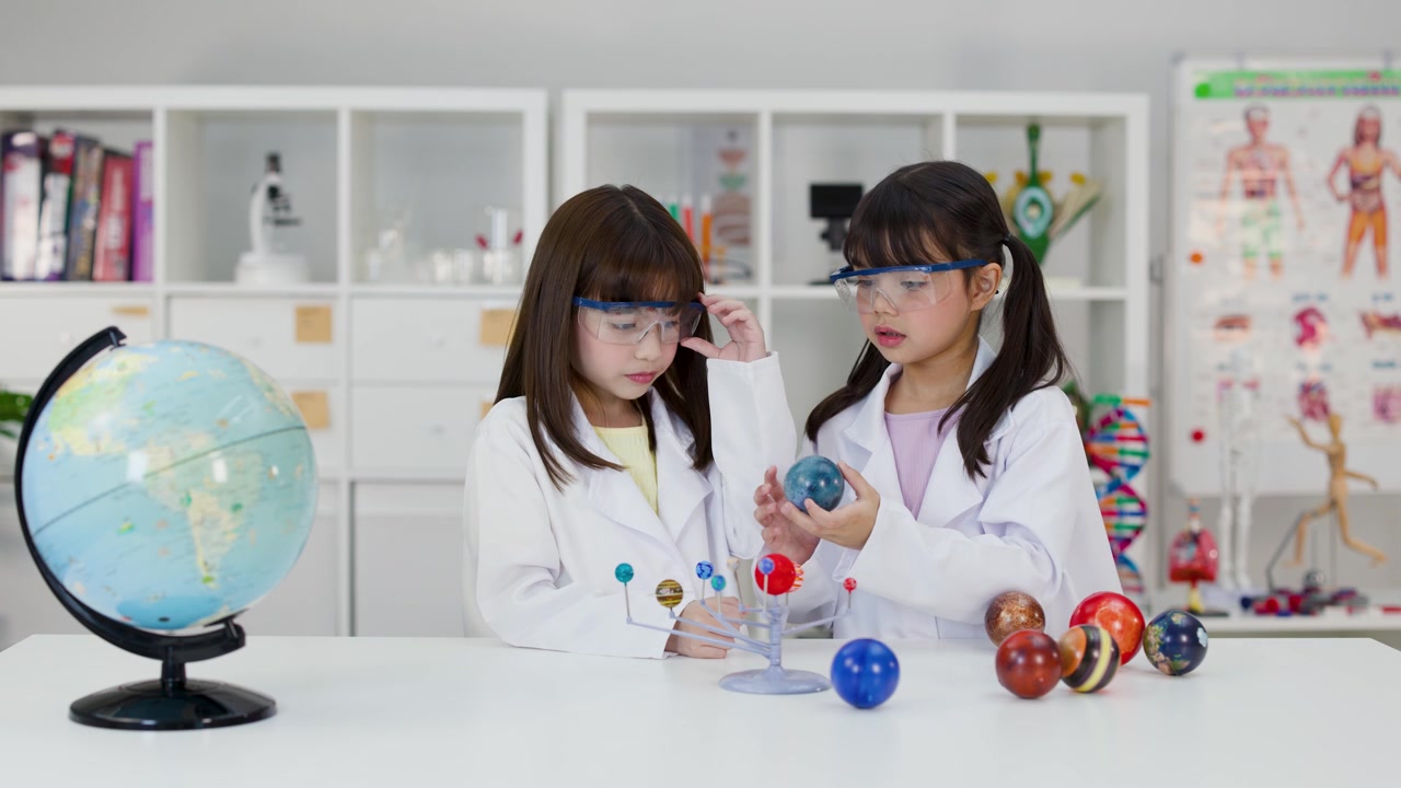 Kaksi tyttöä laboratoriotakeissa ja suojalaseissa tutkii planeettamallia valoisassa luokkahuoneessa.