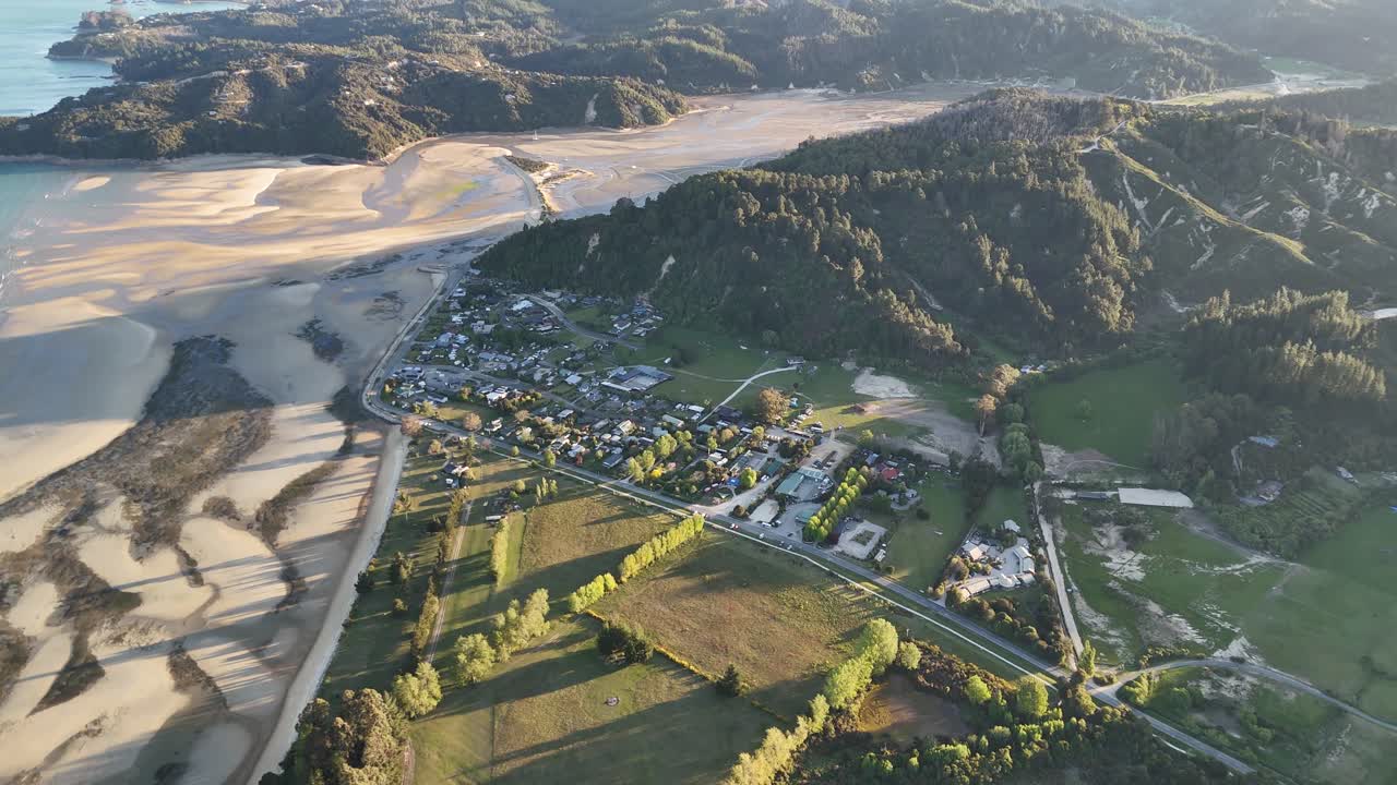 Flygvy vid solnedgång över Marahau i Abel Tasman-regionen, Sydön, Nya Zeeland, som visar kustbebyggelse, landsbygdsjordbruk och tidvattenbankar omgivet av skogsklädda kullar i varmt gyllene ljus.
