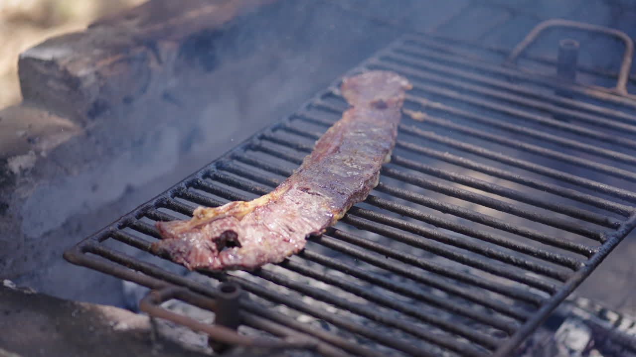 Close up of bovine skirt steak entraña grilling over hot embers on a classic Argentine parrilla, visible smoke, rustic grill bars, rendered fat and browning meat, wood-fired barbecue, outdoor cooking.