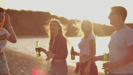 three hot girls are dancing in short t-shirts and shorts with beer on the sand beach at sunset.