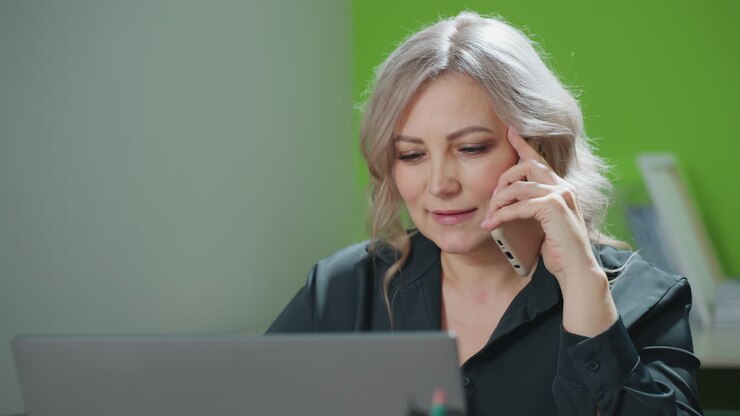 CEO engages in business conversation over phone while simultaneously working on laptop in bright office environment, showcasing leadership, multitasking, and professional communication skills