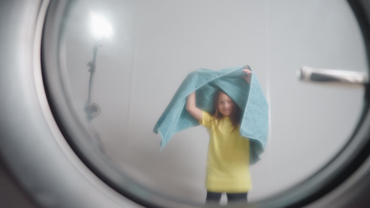 Washer door window shows playful child holding blue towel, covering head and twirling with joy, movement visible through glass, laundry setting, fun moment, clean textiles, modern laundromat vibe