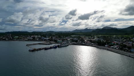 dramatic, establishing aerial view of a lone island's harbor and rural community in background with high contrast clouds and mountains