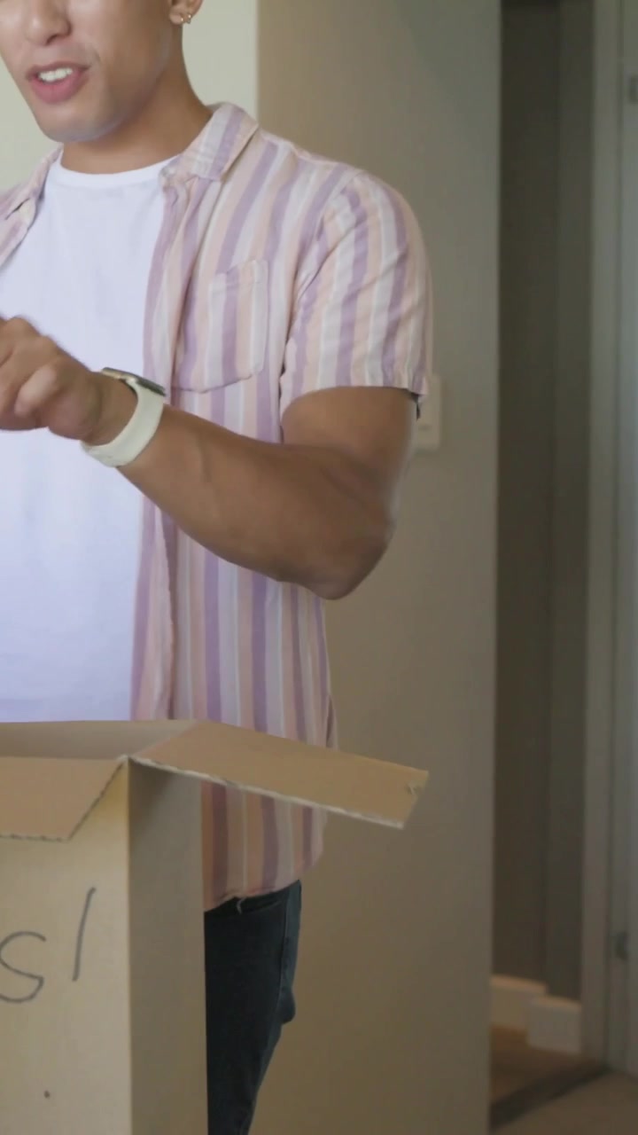 Vertical video: Returning with two shirts, man in pink shirt packing into open box at home, smiling