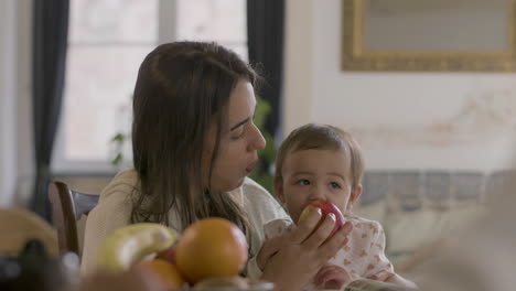 feliz mujer hermosa sentada en la mesa y sosteniendo a su hija mientras comía una manzana juntos en la mañana