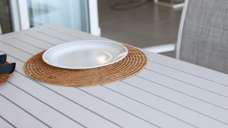 A person arranges a table setting with a plate and cutlery on a woven mat in a bright room