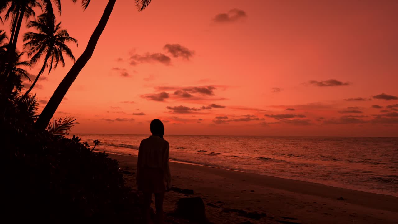A breathtaking view captures a lone woman strolling along a pristine, palm-lined beach in Fuvahmulah, Maldives at sunset.