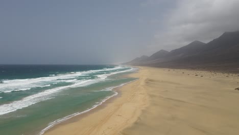 toma aérea de grandes olas rompiendo en la larga playa blanca de cofete con hermosas montañas en el fondo en fuerteventura, isla canaria