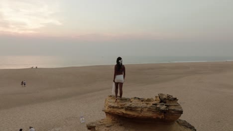 aerial orbit shot of a woman standing on a coastal rock, gazing at the misty ocean and wide sandy beach