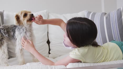 Caucasian-girl-spending-time-at-home,-lying-on-sofa-with-face-mask,-playing-with-pet-dog,-in-slow-mo