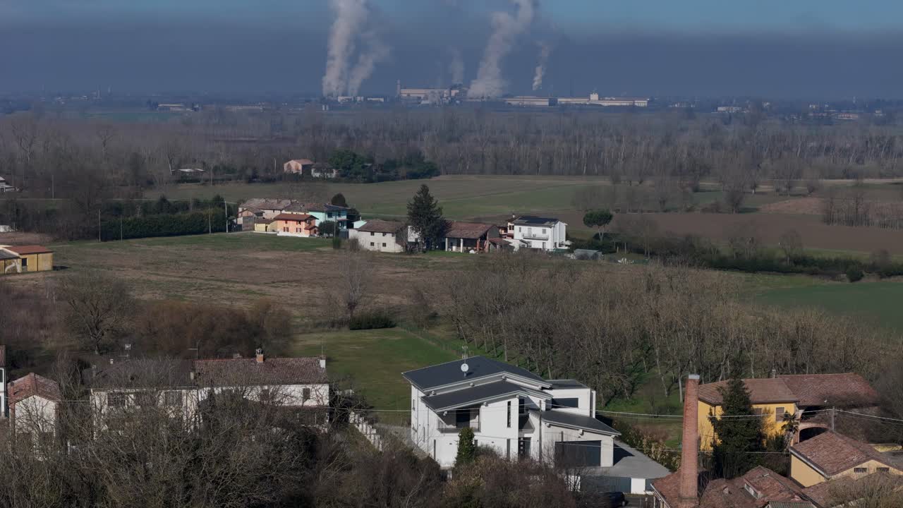 Drone reveals factory chimneys emitting smoke in flat industrial zone—set against forested foreground and clear sky, highlighting environmental tension and atmospheric contrast