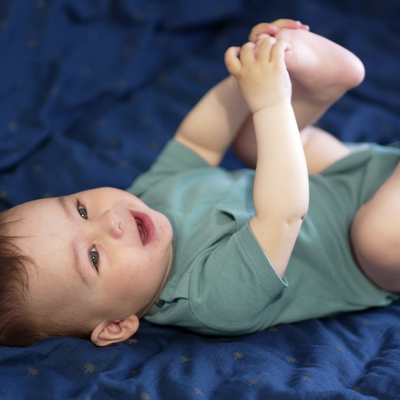 Healthy beautiful kid playing with his feet holding them up. Adorable child laughing cheerfully. Close up. Blue backdrop