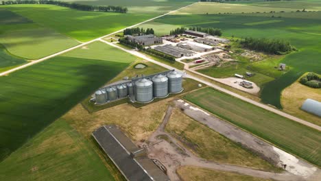 Vista-Aérea-De-La-Torre-De-Silos-Agrícolas-De-Almacenamiento-De-Productos-Agrícolas-Y-Elevadores-De-Granos-En-Medio-De-Un-Campo-Cultivado-Verde,-Alejar