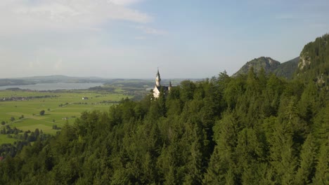 aerial establishing shot of neuschwanstein castle hidden behind alpine trees on summer day