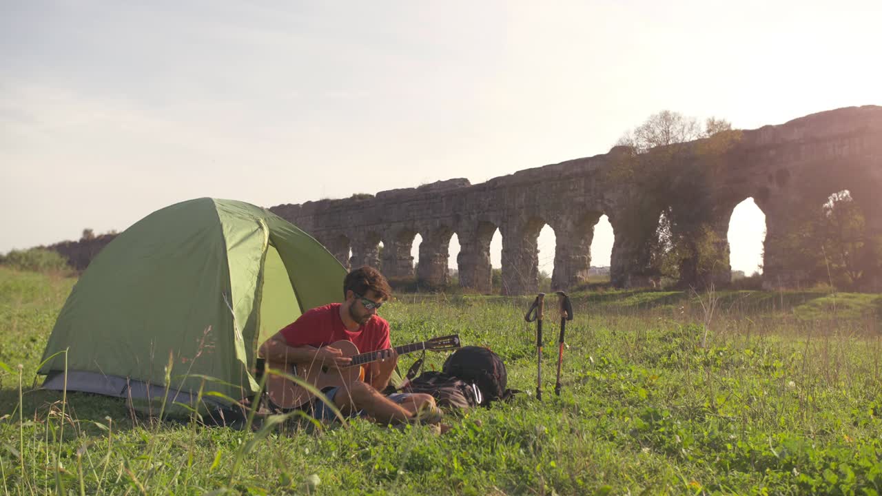 캠핑 텐트 밖에서 기타를 연주하는 젊은 배 타는 남자 로마 수로 앞에 잔디에 앉아 parco degli acquedotti의 공원 유적에서 로마에서 해가 지는 동안 땅 속 느린 동작에 을
