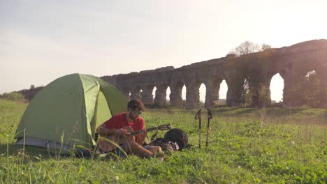 young man backpacker playing guitar outside camping tent sitting on grass in front of roman aqueduct arches in parco degli acquedotti park ruins in rome at sunset sticks in ground slow motion