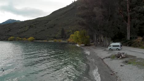 slowmo - motorhome by beautiful blue lake wakatipu, queenstown, new zealand with mountains fresh snow cloudy sky in background - aerial drone