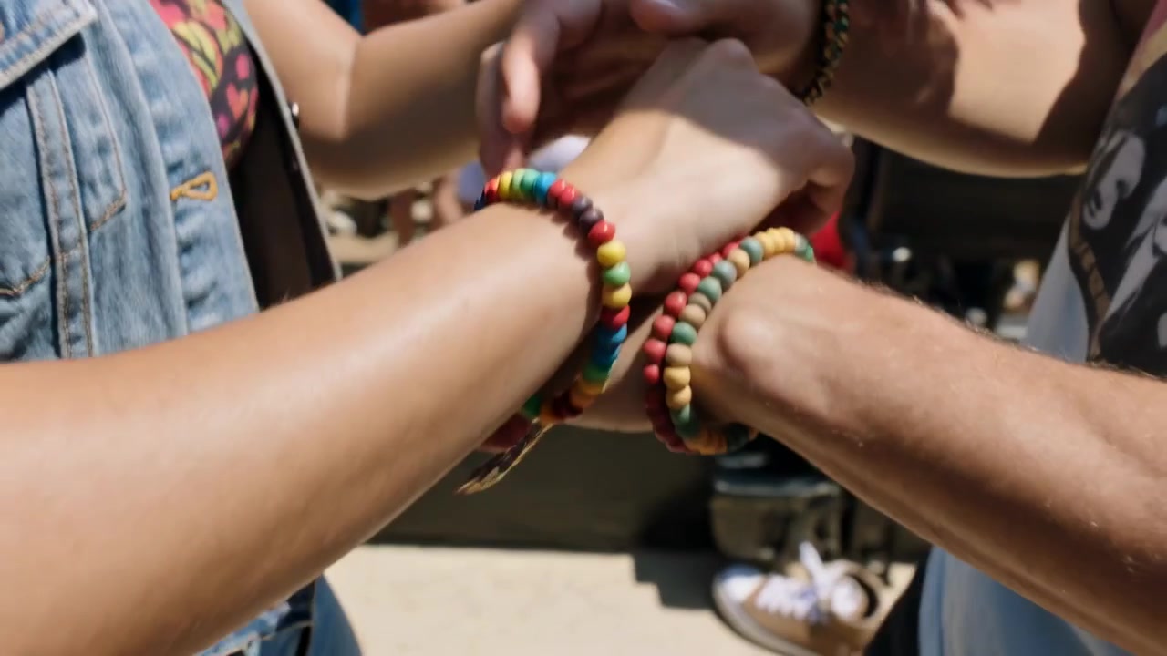 Donna che ride con gilet di jeans, appoggiata a un uomo con maglietta stampata, mentre si scambiano braccialetti da amici a un festival.