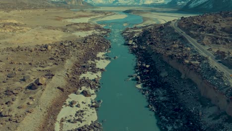 Drone-is-flying-slowly-backwards,-over-a-river-in-Skardu-with-a-view-of-a-high-mountain-range-covered-in-snow