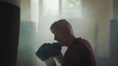 happy man punches heavy bag fast in sports center. smiling sportsman beats equipment by arms in foggy gym slow motion. combat style improvement