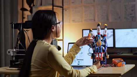 back view of teen asian girl with 3d printing checking a cyborg hand while designing it on a laptop at home